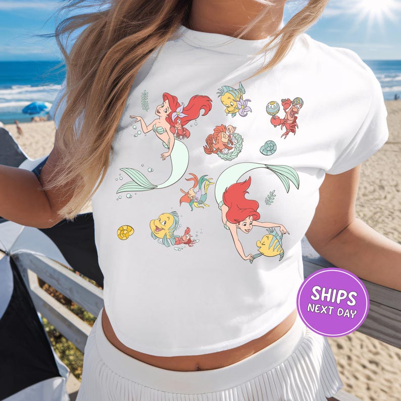 A woman wearing a white t-shirt with a design featuring cartoon mermaid characters.