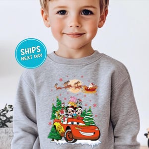 A young boy wearing a gray sweatshirt with a Christmas-themed design featuring characters from the Disney movie Cars and a sleigh ride.