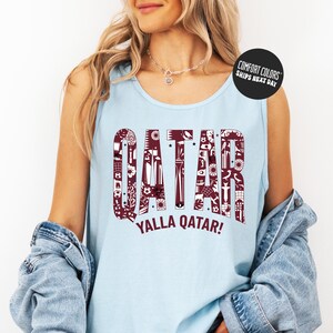 A woman wearing a light blue tank top with the word "QATAR" printed on it, along with the phrase "YALLA QATAR!" printed below. She is also wearing a denim jacket and has a necklace around her neck.