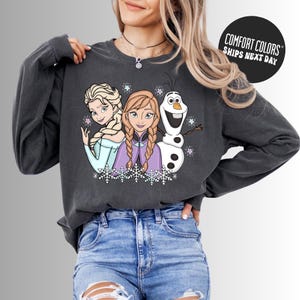 A woman wearing a gray sweatshirt with a graphic of three cartoon characters - two girls and a snowman - smiling and posing together.