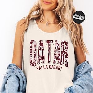 A woman wearing a white tank top with the word "QATAR" printed on it, along with the phrase "YALLA QATAR!" printed below. She is also wearing a denim jacket and has her hands in her pockets.