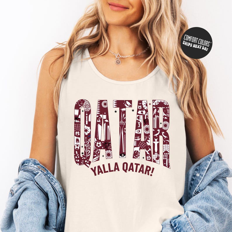 A woman wearing a white tank top with the word "QATAR" printed on it, along with the phrase "YALLA QATAR!" printed below. She is also wearing a denim jacket and has her hands in her pockets.