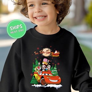A young child wearing a black sweater with a Christmas-themed design featuring a red car, Santa Claus, and other holiday characters.