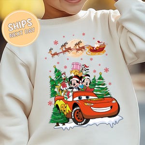 A white sweater with a Christmas-themed design featuring characters from the Disney movie Cars and a Santa Claus sleigh in the background.