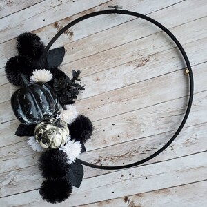 May include: A black metal wreath with white and black faux pumpkins and black and white pom poms. The wreath is on a white wood background.