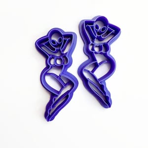 May include: Two purple plastic cookie cutters in the shape of a stylized alien woman. The alien is posed with arms behind the head, in a relaxed position. The cutters are on a white background.