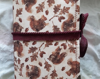 May include: A fabric-covered journal featuring a repeating pattern of brown squirrels and oak leaves on a cream background. The journal is bound with a burgundy cord and has decorative corner protectors. A tassel with a squirrel charm hangs from the bottom.