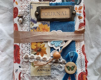 May include: A handmade journal with a pumpkin-themed cover. The journal features a mix of fabrics, lace, and embellishments, including a blue velvet ribbon and a wooden button. The word "JOURNAL" is displayed on a metal label.