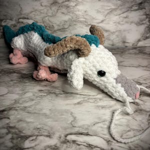 May include: A handmade crochet dragon in white, teal, pink, brown, and gray. The dragon has a white body with teal accents on its back, pink feet, brown horns, and a gray snout. The dragon has black eyes and a white string tail.