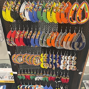 May include: A display rack featuring a variety of handmade beaded earrings in multiple shapes and colors. The collection includes teardrop, circular, and fringed designs crafted with small glass beads in vibrant patterns, including solid colors and multi-colored geometric arrangements.
