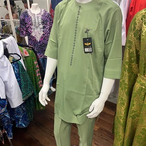 May include: A sage green two-piece outfit consisting of a long-sleeved tunic and matching trousers. The tunic features a round neckline, a vertical row of black geometric embroidery down the center, and a small chest pocket with a black brand tag.