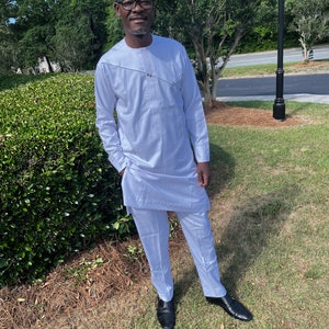 May include: A white long-sleeved tunic shirt with a round neckline and a diagonal decorative silver-toned chain detail across the chest, paired with matching white trousers and black dress shoes.