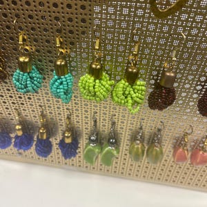 May include: Assortment of colorful beaded earrings displayed on a gold grid. The earrings are made with small beads in various colors, including blue, green, red, and pink.