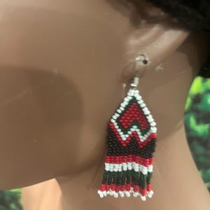May include: A dangle earring crafted from small seed beads in a geometric pattern. The design features a triangular top section in red, white, and dark green, transitioning into a rectangular base with horizontal stripes and a fringed bottom edge.