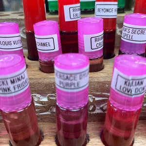 May include: A close-up of a wooden shelf with rows of small pink bottles filled with red liquid. Each bottle has a white label with a different name printed on it, such as "Louis Vuitton", "Chanel", "Versace Diva Purple", "Rihanna Crush", "Nicki Minaj Pink Friday", "Killian Love Don't Be Shy", and "Victoria's Secret Spell".