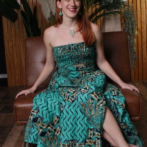 May include: A woman wearing a strapless green and gold patterned maxi skirt with a matching top. The skirt has a high slit on the left side. She is sitting on a brown leather couch.
