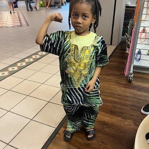 May include: A young boy wearing a green and black patterned shirt and trousers with gold embroidery. He is flexing his bicep and smiling.