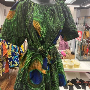 May include: A green dress with a peacock feather pattern, displayed on a mannequin. The dress features a tied waist, short sleeves, and an off-the-shoulder neckline. The fabric has green, blue, and orange accents.