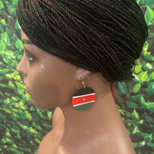 May include: A pair of circular dangle earrings featuring horizontal stripes of black, white, red, and green. The earrings are suspended from gold-toned metal hooks and are displayed on a mannequin head with dark brown braided hair.