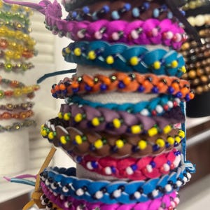 May include: A stack of colorful braided bracelets. The bracelets are in various colors, including blue, pink, orange, and brown, and are adorned with small beads. The bracelets are stacked on a white cylindrical base.