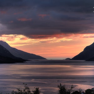 May include: A scenic view of a body of water with mountains on either side. The sky is a vibrant orange and pink, with clouds casting shadows on the water. The mountains are silhouetted against the sky.