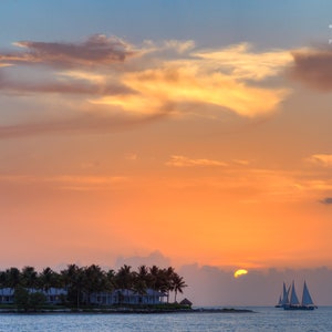 May include: A scenic sunset over a tropical island with palm trees and a sailboat on the water. The sky is a vibrant orange and pink with clouds.
