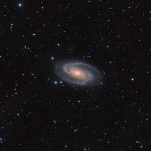 May include: A photograph of a dark night sky with a faint, swirling galaxy in the center. The galaxy is a pale, milky white with a bright, reddish core. There are many stars scattered across the sky, some of which are brighter than others. The image is taken from a dark sky location, so the stars are very visible.