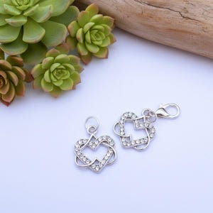 May include: Two silver heart-shaped charms, each with clear crystal detailing. One charm has a lobster clasp. The charms are on a white surface, with green succulents and a piece of wood in the background.