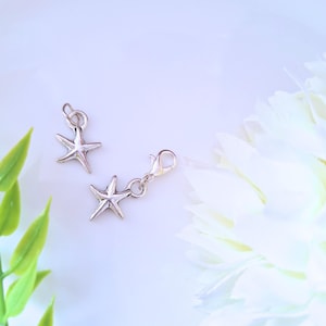 Può includere: Due ciondoli a forma di stella marina color argento. Uno ha un piccolo anello, l'altro un moschettone. I ciondoli sono su sfondo bianco con un fiore bianco e foglie verdi.