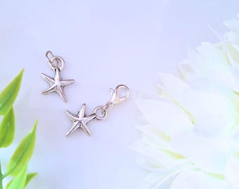 May include: Two silver-colored starfish charms. One charm has a small loop, and the other has a lobster clasp. The charms are on a white background with a white flower and green leaves.
