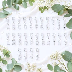 May include: Silver-colored alphabet charms, each with a lobster clasp, are arranged on a white fabric. The letters A through Z are displayed, with each charm approximately 1 inch in height. Green foliage and white flowers surround the charms.