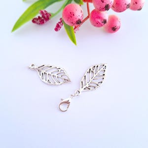 May include: Two silver-colored leaf charms. One charm is a simple leaf shape, and the other has a lobster clasp attached. The charms are on a white surface, with pink berries and green leaves in the background.