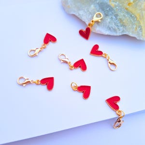 May include: A collection of red heart-shaped charms with gold-coloured clasps, scattered on a white surface. The charms have a glossy finish and a textured border. The clasps are open, ready to attach to a necklace or bracelet.