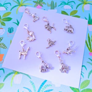 May include: A selection of silver charms with animal designs, such as a teddy bear, lion, fox, giraffe, elephant, dinosaur, and bee. Each charm has a lobster clasp for attaching to bracelets or necklaces. The charms are presented on a white surface.