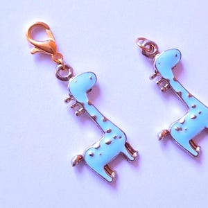 Op de afbeelding: Twee lichtblauwe girafbedels met goudkleurige accenten. Eén bedel heeft een karabijnhaaksluiting, de andere een kleine lus. De giraffen hebben bruine vlekken en staan rechtop. Deze bedels zijn waarschijnlijk voor het maken van sieraden of knutselen.