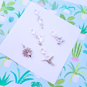 May include: A collection of silver-colored charms featuring various animal designs. The charms include a peacock, a hummingbird, a parrot, a dove, and a swan. Each charm has a lobster clasp for easy attachment. The charms are displayed on a white surface.
