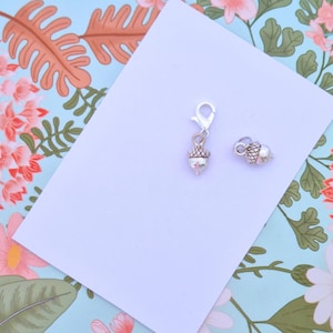 May include: Two silver-coloured acorn charms. One charm has a lobster clasp, and the other is a standalone acorn. The charms are on a white surface, set against a floral patterned background with pink and orange flowers and green leaves.