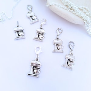 May include: Silver-colored charm pendants in the shape of stand mixers with lobster claw clasps. The charms are scattered on a white surface, with a silver chain and a decorative element in the background.