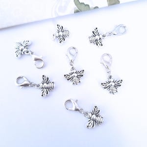 Puede incluir: Varios dijes plateados con forma de abeja con cierres de mosquetón. Los dijes están detallados con alas y segmentos corporales. Los cierres están abiertos, listos para sujetarse a un collar, pulsera u otra joya.