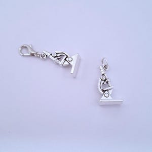 Puede incluir: Dos colgantes plateados con forma de microscopio. Un colgante tiene un cierre de mosquetón. El otro colgante tiene un pequeño lazo en la parte superior. Ambos colgantes están detallados con los componentes de un microscopio.