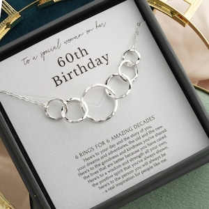 May include: A silver necklace with six interconnected rings, presented in a gift box. The card inside reads "60th Birthday" and includes a message about the recipient's life. The necklace is a thoughtful gift.