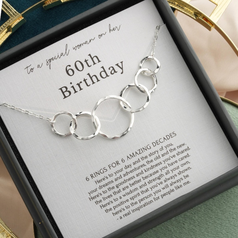 May include: A silver necklace with six interconnected rings, presented in a gift box. The card inside reads "60th Birthday" and includes a message about the recipient's life. The necklace is a thoughtful gift.