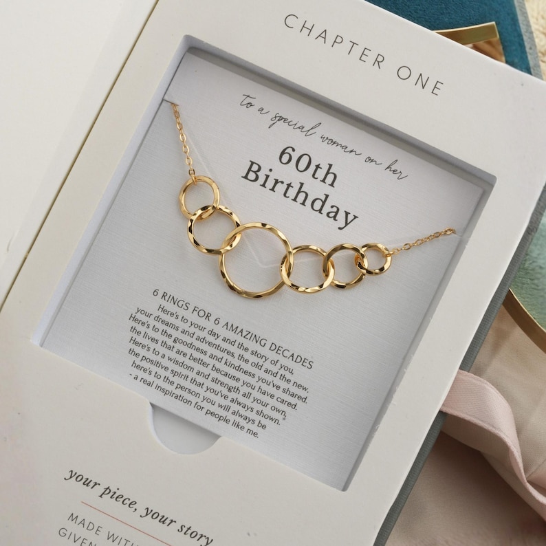 May include: Gold-toned necklace with six interlocking rings, presented in a white gift box. The box features text that reads "60th Birthday" and "6 Rings for 6 Amazing Decades."
