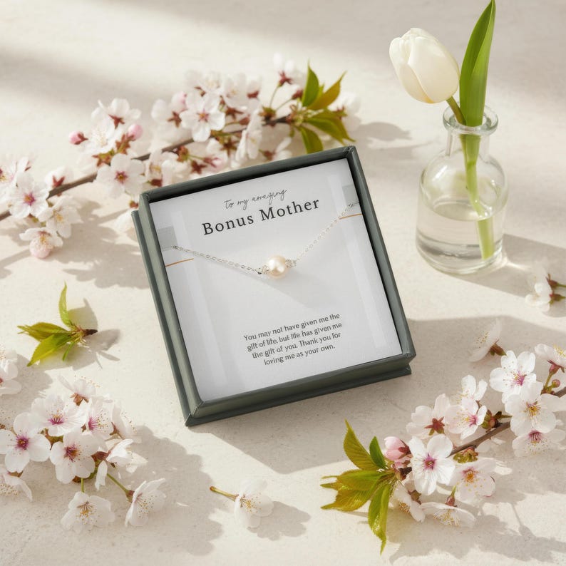 May include: A silver necklace with a pearl pendant is displayed in a gray gift box. The box is surrounded by white and pink cherry blossoms and a small glass vase with a white tulip. The text on the box reads "To my amazing Bonus Mother."