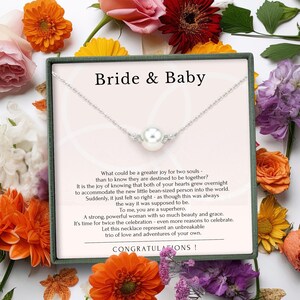 May include: A silver necklace with a large pearl pendant and small accent stones, presented in a gift box with the words "Bride & Baby." The box is surrounded by colorful flowers, with text that reads "Congratulations!"