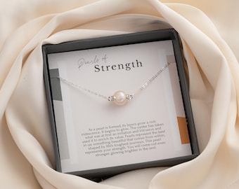 Puede incluir: Un delicado collar con un colgante de perla única, presentado en una caja de regalo. La tarjeta interior dice "Perlas de Fuerza" e incluye texto inspirador sobre perlas. El collar está sobre una tela suave de color crema.