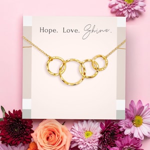 Op de afbeelding: Een goudkleurige ketting met vijf in elkaar grijpende cirkels, gepresenteerd op een lichtroze kaart met de woorden "Hope. Love. Shine." De kaart is omgeven door roze en paarse bloemen, wat een vrouwelijke en elegante uitstraling creëert.