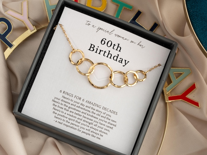 May include: Gold-toned necklace with six interlocking rings, presented in a gift box. The card inside reads "60th Birthday" and includes a message about celebrating six decades. The necklace is a thoughtful gift.