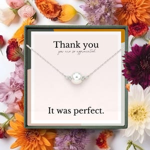 Może przedstawiać: Srebrny naszyjnik z dużą białą perłą, zapakowany w pudełko prezentowe z napisem "Thank you, you are so appreciated" i "It was perfect." Pudełko otoczone jest kolorowymi kwiatami.