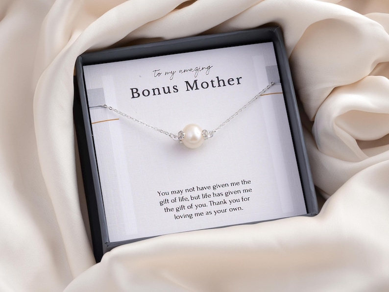 May include: A silver necklace with a single pearl pendant, presented in a black gift box. The box contains a card with the text "To my amazing Bonus Mother" and a heartfelt message. The background is a soft, cream-colored fabric.